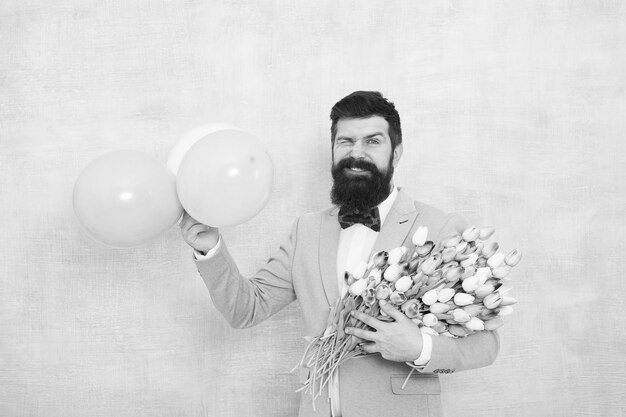Dostawa kwiatów Romantyczna randka dla dżentelmena Życzenia urodzinowe Pewność siebie i charyzma Mężczyzna brodaty dżentelmen muszka trzyma balony i bukiet Pan robi dla niej romantyczną niespodziankę