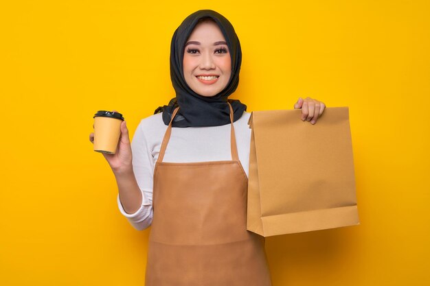 Dostawa młoda Azjatycka kobieta barista w białej koszulce fartuch praca jako sprzedawca usługi kurierskie przytrzymaj brązowy wyczyść pusty papier rzemieślniczy torba na wynos makiety i filiżanka kawy na żółtym tle Uruchomienie firmy