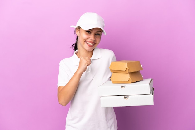 Dostawa pizzy Rosyjska dziewczyna w mundurze roboczym zbierając pudełka po pizzy i hamburgery na fioletowym tle świętująca zwycięstwo