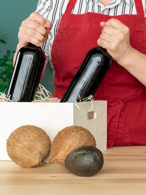 Dostawa w czerwonym fartuchu Wkłada butelki wina do białego pudełka z kokosami i awokado, koncepcja zamówienia online.