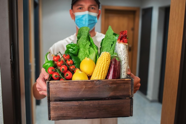 Dostawca w masce ochronnej trzymający przy wejściu papierową torbę z jedzeniem Kurier wręcza klientowi pudełko ze świeżymi warzywami i owocami
