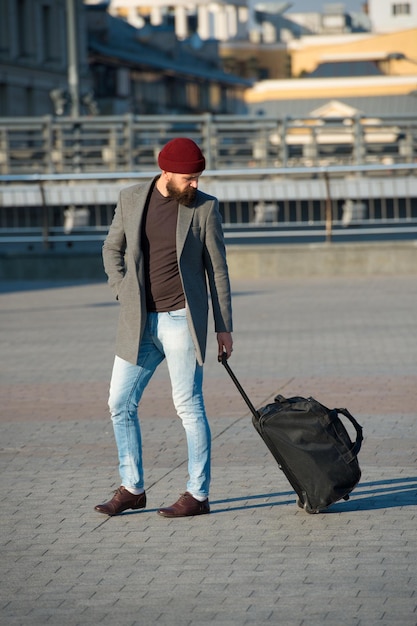 Dostosuj życie w nowym mieście Podróżnik z walizką przylot na lotnisko stacja kolejowa tło miejskie Hipster gotowy cieszyć się podróżą Noś torbę podróżną Mężczyzna brodaty hipster podróżuje z torbą bagażową na kółkach