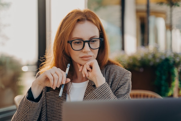 Doświadczona bizneswoman z rudymi włosami ubrana w eleganckie ubrania ogląda film instruktażowy o tworzeniu programu startowego robi notatki zapisuje ważne informacje trzyma uważnie piórem patrzy na ekran