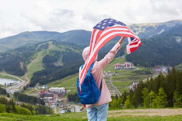 Doświadczony Podróżnik Ubrany W Dżinsy, Różową Kurtkę Z Kapturem I Niebieski Plecak, Podnoszący Dużą Flagę Usa, Poczucie Wolności, Podróż Po Mieście