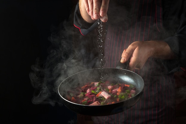 Doświadczony szef kuchni soli warzywa na gorącej patelni Koncepcja menu dla hotelu na czarnym tle