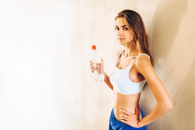 Dosyć sporty dziewczyna stoi blisko ściany po trenować z butelką woda.