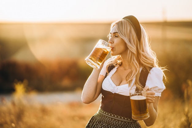 Dosyć szczęśliwa blondynka w dirndl, tradycyjnej festiwalowej sukni, trzyma dwa kufle piwa outdoors w polu