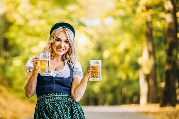 Dosyć szczęśliwa blondynka w dirndl, tradycyjnej festiwalowej sukni, trzymająca dwa kufle piwa na zewnątrz w lesie