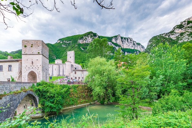 Dramatyczne niebo nad średniowieczną wioską nad rzeką, San Vittore Frasassi, Marche, Włochy. Romantyczne niebo i chmury nad krajobrazem gór, cel turystyczny.