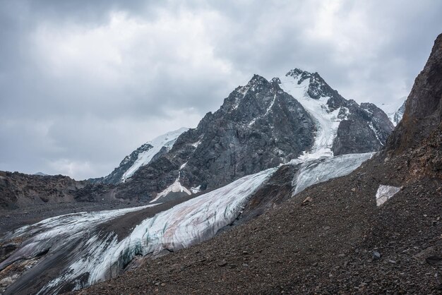 Dramatyczny krajobraz z wiszącym lodowcem na tle dużej śnieżnej góry z ostrym skalistym szczytem pod szarym pochmurnym niebem Lodowiec z lodowym gzymsem na dużej wysokości Ponura sceneria w górach w pochmurny dzień