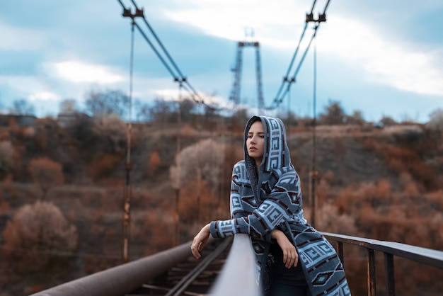 Dramatyczny portret młodej nastoletniej dziewczyny z kapturem stojącej na opuszczonym starym zardzewiałym moście z perspektywiczną powierzchnią.