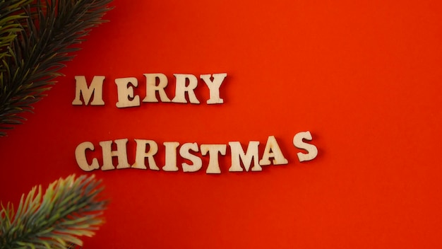 Drewniane litery na ciemnoczerwonym tle napisane po angielsku „Wesołych Świąt” obok zielonej gałęzi jodły
