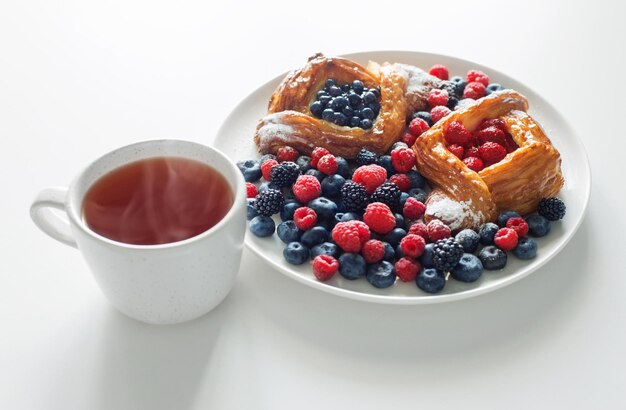 Duński z malinami i jagodami z filiżanką herbaty na białym stole
