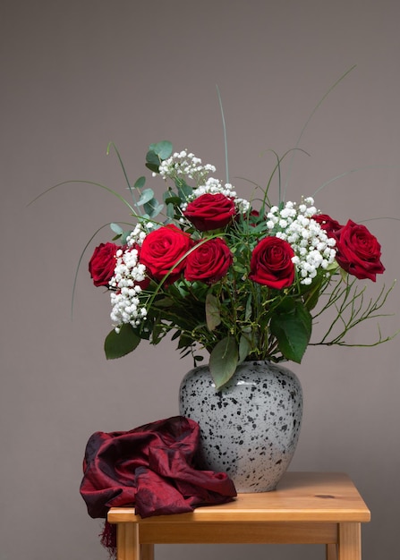 Duży bukiet czerwonych róż i białych kwiatów w szarym wazonie na drewnianym stole obok bordowego szalika Szare tło Martwa natura