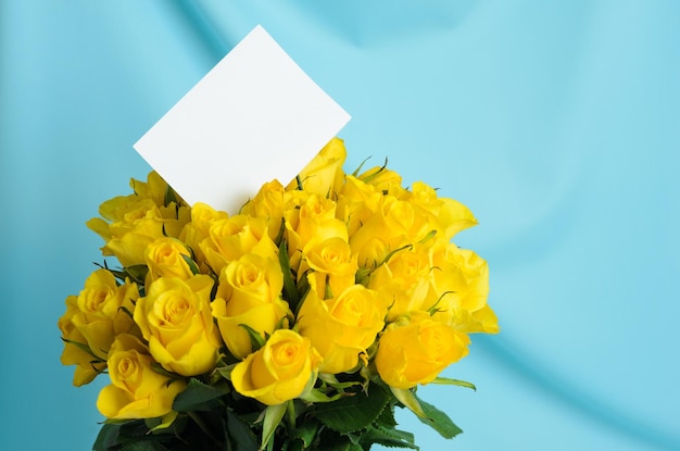 Duży bukiet delikatnych żółtych róż z białą kartką z życzeniami na niebieskim falistym tle