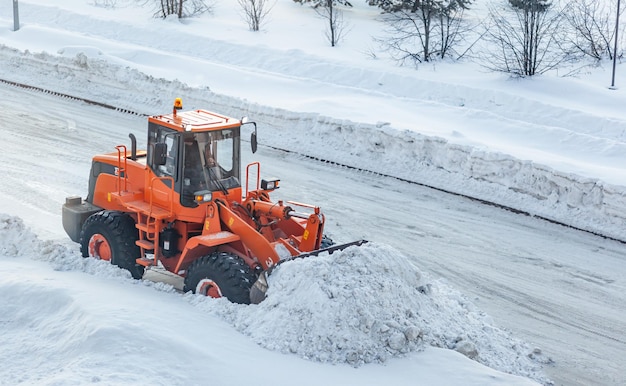 Duży pomarańczowy traktor usuwa śnieg z drogi i ładuje go na ciężarówkę. Sprzątanie i odśnieżanie dróg w mieście zimą
