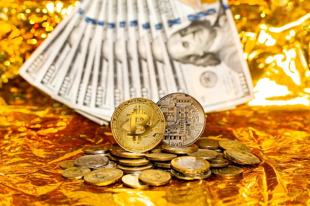 Dwa bitcoiny na stosie monet na tle studolarowych banknotów ułożonych w wachlarz na złotym tle.