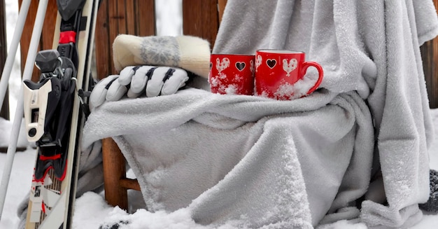 Dwa czerwone kubki z sercem na kratce postawione na krześle w śniegu i sprzęcie narciarskim na tarasie