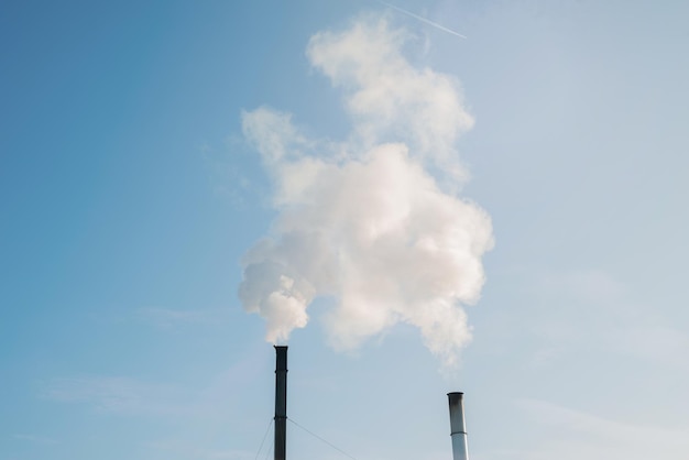 Dwa dymiące kominy fabryczne i ślad samolotu na tle błękitnego nieba. Zwiększenie emisji CO2 i gazów cieplarnianych.