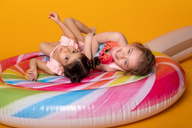 Dwa dziecko dziewczynka w kostiumie kąpielowym leżąc zabawy na kolorowym dmuchanym materacu lizak na żółtej powierzchni.