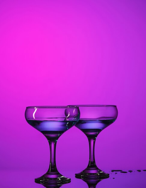 Dwa kieliszki do wina nadziewane stojące na stole w studio. Żywe, jasne, kolorowe oświetlenie. Modna w 2018 roku żarówka ultrafioletowa. Dekoracja artystyczna z odcieniem mistycznego koloru