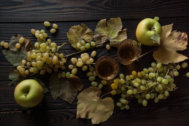 Dwa kryształowe kieliszki z białym winem, jabłkami, winogronami i suchymi liśćmi na drewnianej powierzchni. Widok z góry