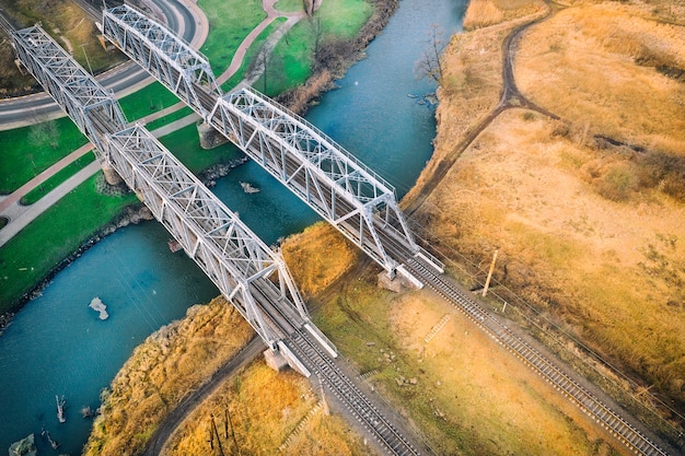 Zdjęcie dwa metalowe mosty, z siecią kolejową, nad małą rzeką, jeden brzeg zielony, z trawnikiem, drugi żółty, z suchą trawą. kontrastujący krajobraz. widok drona.