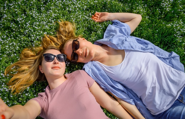 Dwa młoda szczęśliwa kobieta robi selfie na zielonej trawie plenerowej. Kochająca żeńska para kłama twarz w twarz na kwiatonośnej łące i trzyma ręki. Spokój, młodość, miłość unisex, przyjaźń.