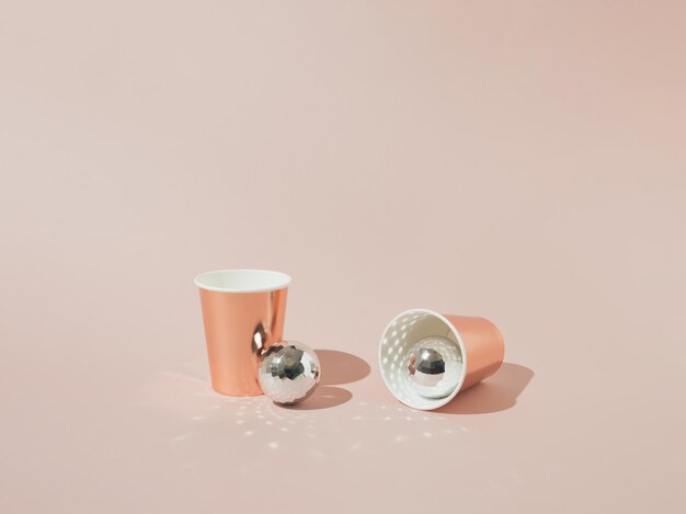Dwa papierowe kubki na napoje w kolorze różowego złota z błyszczącymi kulkami dyskotekowymi na różowym tle. Minimalna koncepcja.
