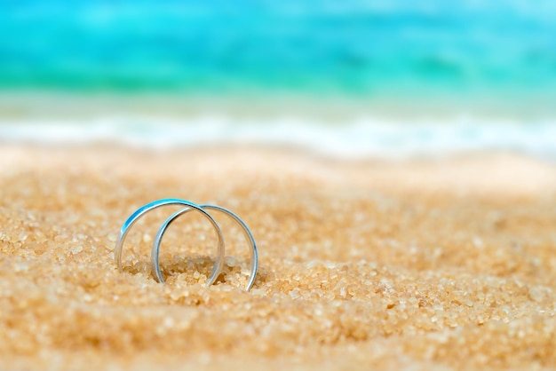 Dwa srebrne obrączki ślubne w piasku na tle plaży i morza