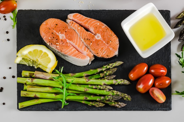 Dwa świeże surowe steki z łososia z warzywami i przyprawami: szparagi, pomidory, pieprzu, rukoli, cytryny i oliwy z oliwek na szarym tle. Pojęcie diety zdrowej żywności.