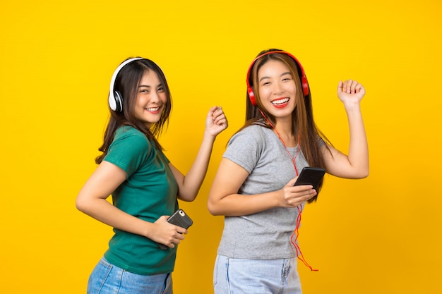 Dwa Szczęścia Azjatycka Uśmiechnięta Młoda Kobieta W Bezprzewodowych Słuchawkach Do Słuchania Muzyki Za Pomocą Inteligentnego Telefonu Komórkowego I Tańca Na Izolowanej żółtej ścianie, Stylu życia I Wypoczynku Z Koncepcją Hobby
