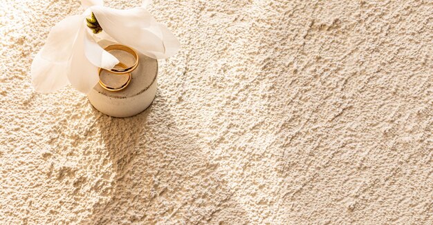 Zdjęcie dwa złote pierścienie ślubne leżą na okrągłym betonowym naturalnym podium z białym kwiatem, wygrawerowanym piaskowym betonowym tłem dla tekstu