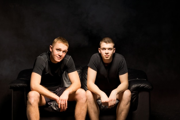 Dwaj Poważnie Wysportowani Młodzi Mężczyźni Siedzący Obok Siebie Na Czarnej Sofie