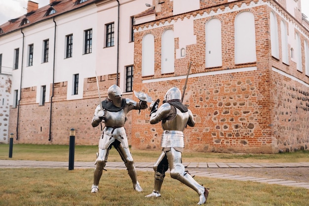 Dwaj rycerze w starożytnej metalowej zbroi stoją pod kamiennym murem zamku walcząc na miecze