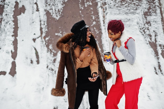 Dwie African American Womans W Kożuch I Futrze Pozowane W Zimowy Dzień Na śnieżnym Tle Z Filiżankami Kawy I Patrząc Na Telefon Komórkowy