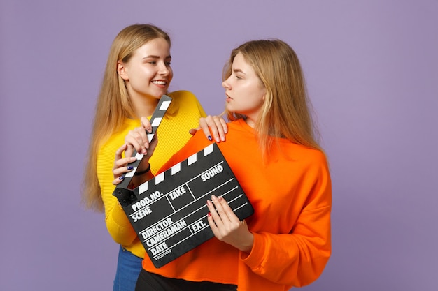 Dwie atrakcyjne młode blond bliźniaczki siostry dziewczyny w kolorowe ubrania trzymają klasyczny czarny film robi clapperboard na białym tle na fioletowej niebieskiej ścianie. Koncepcja życia rodzinnego osób. .