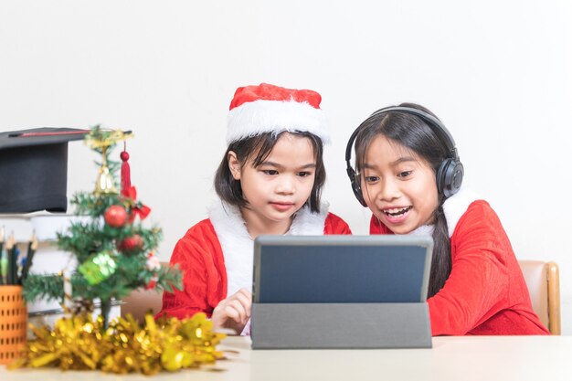 Dwie azjatyckie dzieci małe dziewczynki noszą Świętego Mikołaja na czacie z przyjacielem na cyfrowym tablecie. Boże Narodzenie wakacje koncepcja Stock Photo