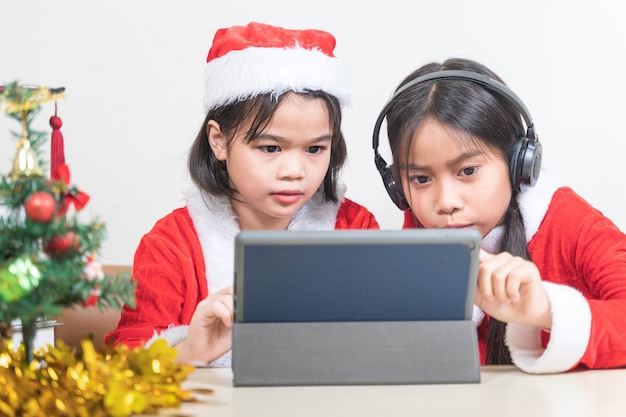Dwie azjatyckie dzieci małe dziewczynki noszą Świętego Mikołaja na czacie z przyjacielem na cyfrowym tablecie. Boże Narodzenie wakacje koncepcja Stock Photo