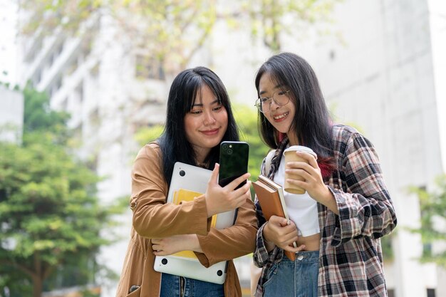 Zdjęcie dwie azjatyckie studentki stoją razem w parku kampusu i patrzą na smartfon.