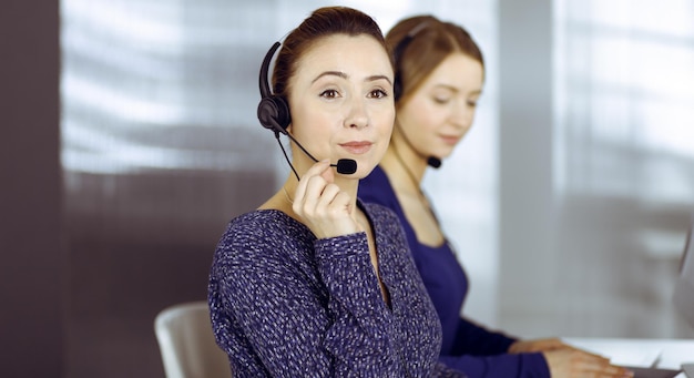 Dwie bizneswoman rozmawiają z klientami przez zestawy słuchawkowe, siedząc przy biurku w nowoczesnym biurze. Zróżnicowana grupa ludzi w call center. Telemarketing i obsługa klienta.