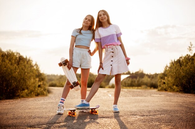 Dwie blond nastolatki w modnych ciuchach z deskorolką w letnim parku na świeżym powietrzu