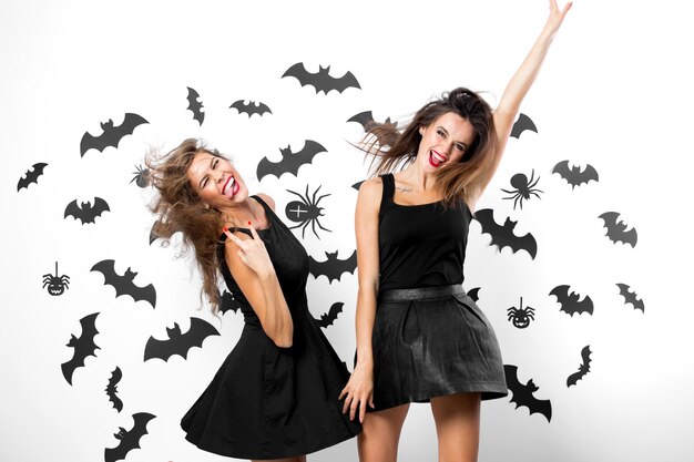 Dwie brunetki w czarnych sukienkach bawią się na tle ściany z nietoperzami i pająkami. Impreza halloween'owa .