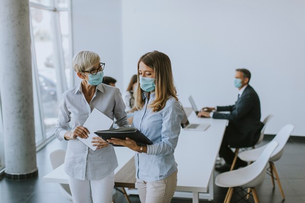 Dwie dojrzałe i młode kobiety biznesu rozmawiają w biurze i noszą maskę chroniącą przed wirusami