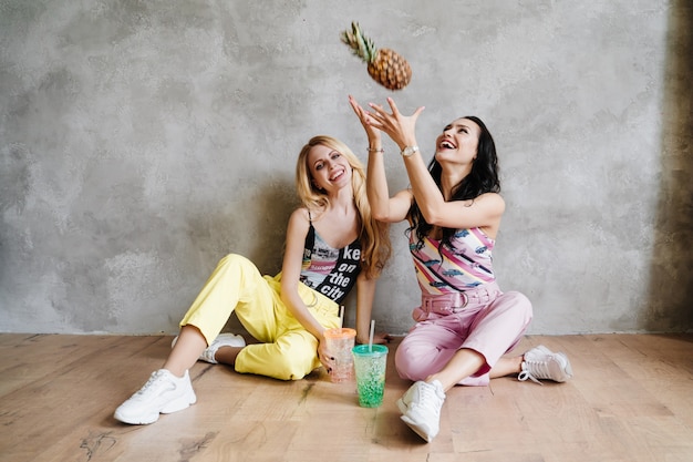 Dwie dziewczyny, brunetka i blondynka, w kolorowych spodniach. Jedz ananasa i pij koktajle.