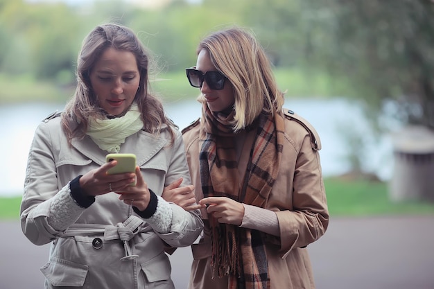 dwie dziewczyny patrzą na telefon jesienny spacer / jesienny płaszcz miejski park, jesienna moda, styl, wygląd, ubrania