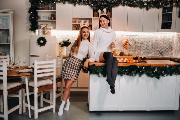 Dwie dziewczyny w przytulnym domowym otoczeniu w kuchni z szampanem w dłoniach na Boże Narodzenie. Uśmiechnięte dziewczyny piją szampana w świąteczny wieczór.