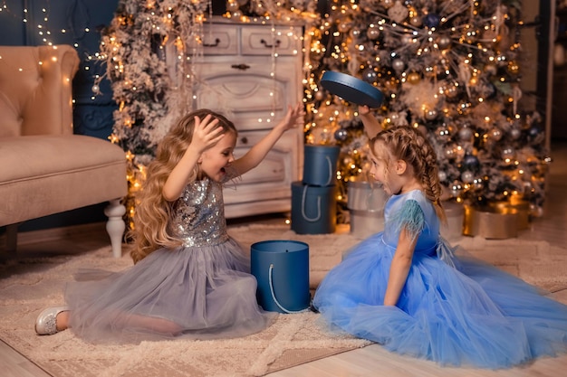 Dwie eleganckie dziewczyny w pięknych sukienkach dają sobie prezent na tle choinki ze światłami