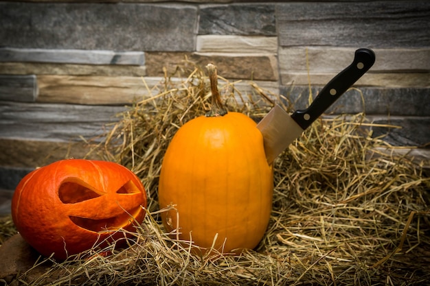 Zdjęcie dwie halloweenowe dynie leżą na sianie na tle kamiennego noża wbitego w żółtą dynię