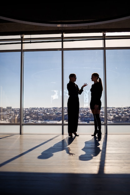 Dwie kobiety biznesu cieszące się widokiem na miasto i rozmawiające stojąc przy dużym oknie w biurze